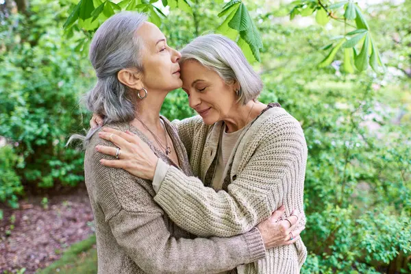 A middle-aged lesbian couple in cardigans hug each other affectionately in a lush green forest. — Stock Photo