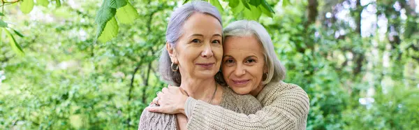 Two middle-aged women in cardigans embrace each other in a lush green forest. — Stock Photo