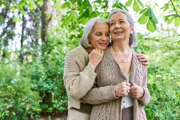 Two middle-aged women wearing cardigans hug each other in a forest setting. — Stock Photo