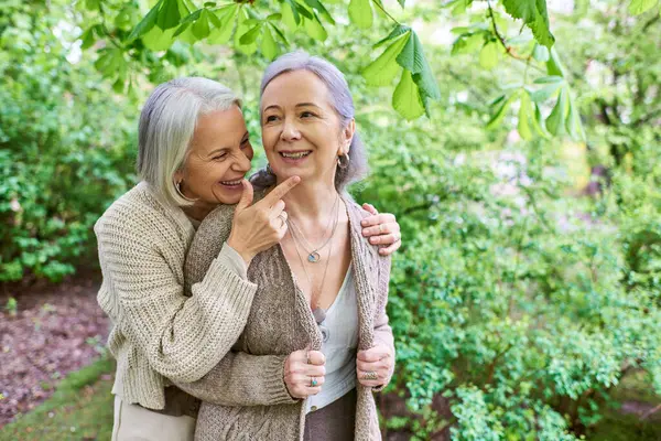 A middle-aged lesbian couple in cardigans, with gray hair, laugh and hug each other in a beautiful green garden setting. — Stock Photo