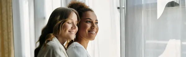 Two women, embracing each other, smile while looking out of a window. — Stock Photo