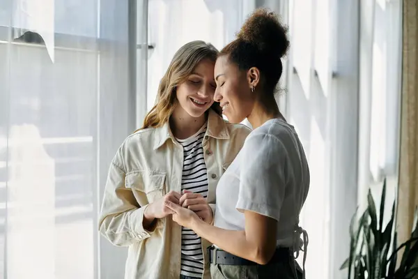 Two women in casual clothing stand close to each other by a window, smiling and holding hands. — Stock Photo