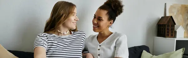 A diverse lesbian couple smiles at each other while sitting on a couch in their living room. — Stock Photo