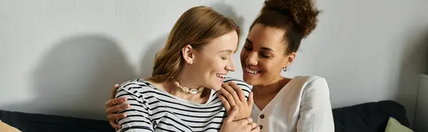 A diverse lesbian couple smiles and hugs each other while sitting on a couch at home. — Stock Photo
