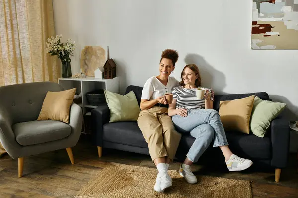 Un couple de lesbiennes diversifié profite d'un après-midi de détente à la maison, partage le café et sourit sur le canapé. — Photo de stock