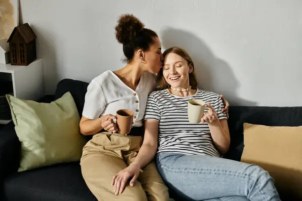 Two women, in casual wear, share a cozy moment on a couch with cups of coffee. — Stock Photo