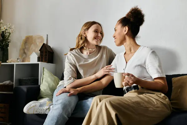 Two women in casual clothing sit on a couch, sharing coffee and a warm conversation at home. — Stock Photo