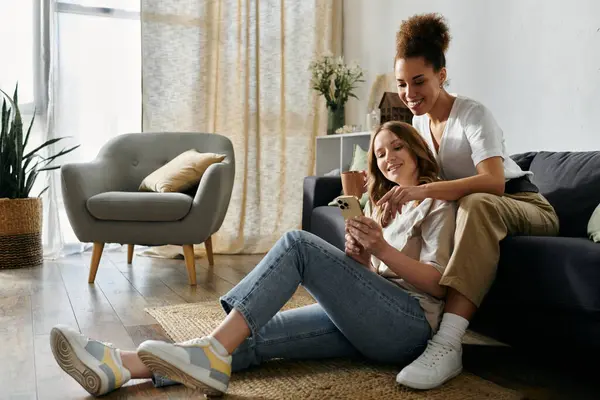 Two women relax at home, one sitting on the floor and the other on a couch, looking at a phone together. — Stock Photo