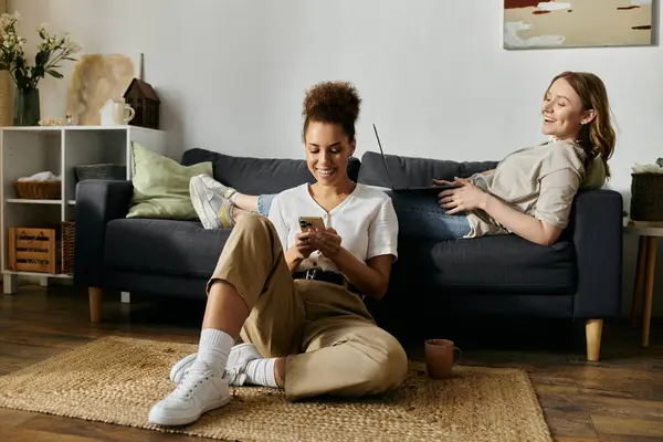 Ein lesbisches Paar genießt einen lockeren Nachmittag zu Hause, wobei die eine ihr Telefon benutzt und die andere es sich auf der Couch gemütlich macht. — Stockfoto