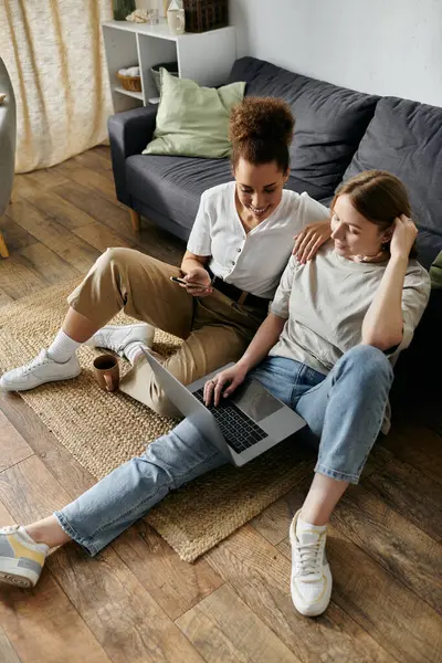 Una coppia lesbica si siede sul pavimento in un soggiorno, uno usando un computer portatile mentre l'altro guarda il suo telefono. — Foto stock