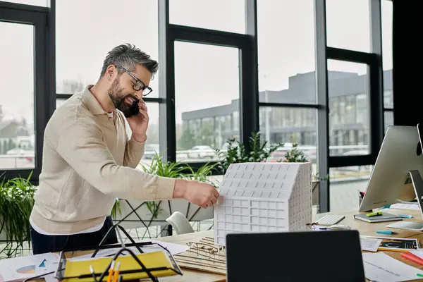 Ein gutaussehender Geschäftsmann mit Bart steht in einem modernen Büro, telefoniert und gestikuliert in Richtung eines weißen Baumodells auf einem Tisch.. — Stockfoto