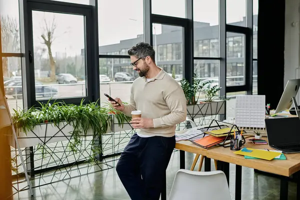 Un homme d'affaires barbu dans un pull beige se tient près d'un bureau dans un bureau moderne, vérifiant son téléphone et tenant une tasse de café. — Photo de stock