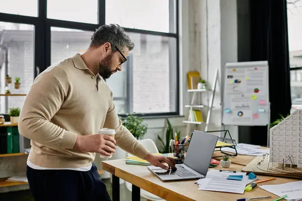 Un hombre de negocios guapo con barba trabaja en su portátil en un espacio de oficina moderno, sosteniendo una taza de café en una mano. — Stock Photo