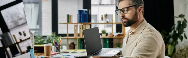 Un hombre de negocios guapo con barba trabaja en su portátil en un entorno de oficina moderno. - foto de stock