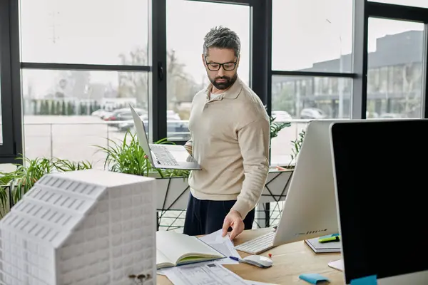 Un homme d'affaires avec une barbe travaille sur un ordinateur portable et examine les plans architecturaux dans un bureau moderne. — Photo de stock