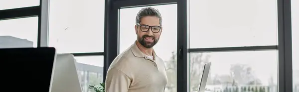 A confident, bearded businessman in glasses stands in a modern office space with a window behind him. — Stock Photo