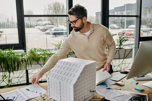 Un bel homme d'affaires barbu en lunettes travaille dans un bureau moderne, examinant un modèle de bâtiment sur son bureau. — Photo de stock