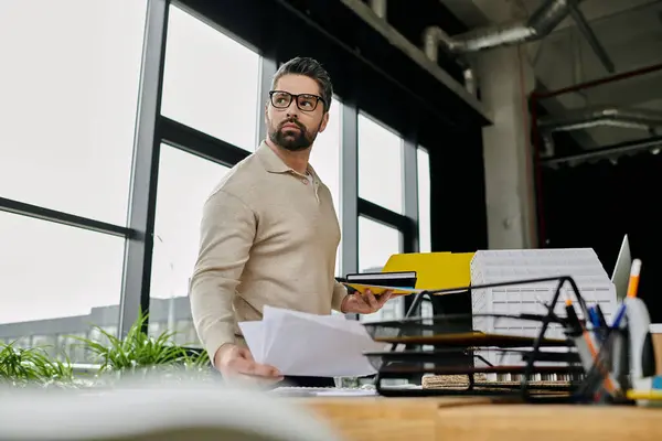 A man with a beard, wearing glasses, stands in a modern office, holding papers and looking towards a window. — Stock Photo