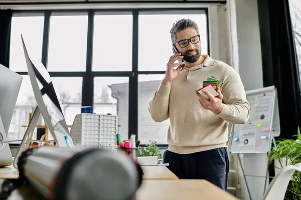 Un bel homme d'affaires avec une barbe se tient dans un bureau moderne, examinant des échantillons colorés tout en parlant sur son téléphone portable. — Photo de stock
