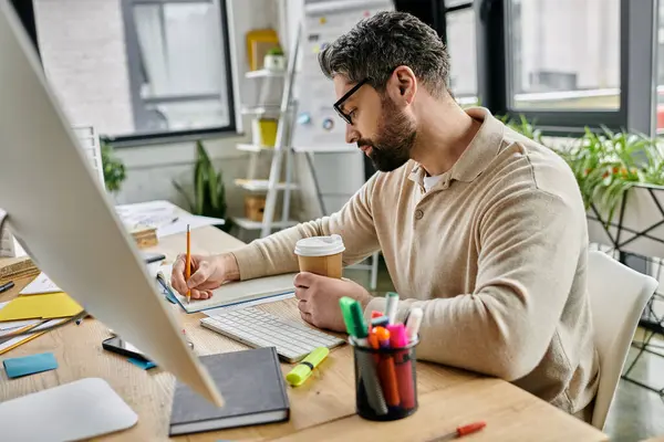 Ein gutaussehender Geschäftsmann mit Bart sitzt an seinem Schreibtisch in einem modernen Büro und schreibt nachdenklich in ein Notizbuch.. — Stockfoto