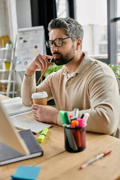 Un bell'uomo d'affari con la barba medita su un progetto mentre siede alla sua scrivania in un ufficio moderno. Sta tenendo una tazza di caffè in una mano e una matita nell'altra. — Foto stock