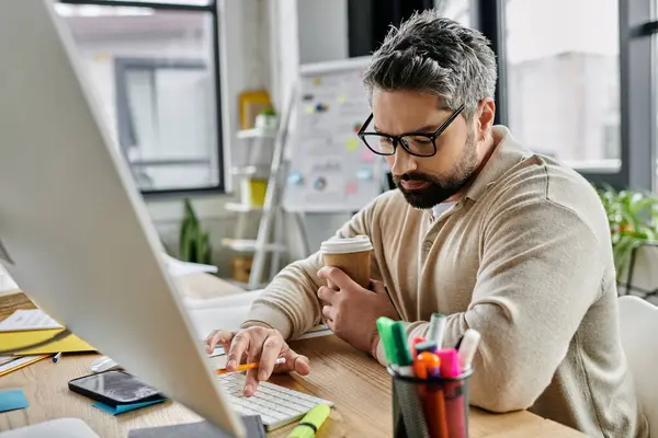 Un bell'uomo d'affari con la barba si siede alla scrivania in un ufficio moderno, digitando su una tastiera del computer mentre tiene in mano una tazza di caffè. — Foto stock