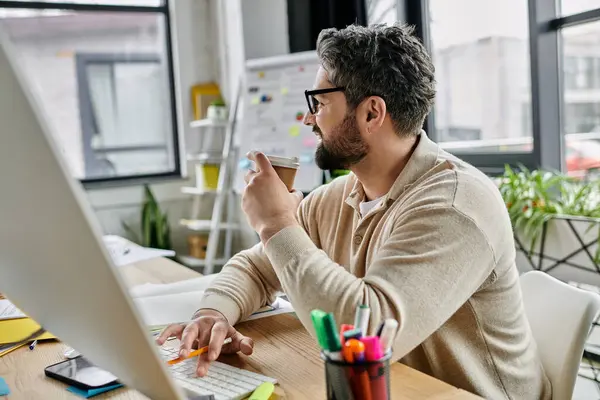 Un bell'uomo d'affari con la barba si prende una pausa dal suo lavoro, sorseggiando un caffè e digitando su un computer in un ufficio moderno. — Foto stock