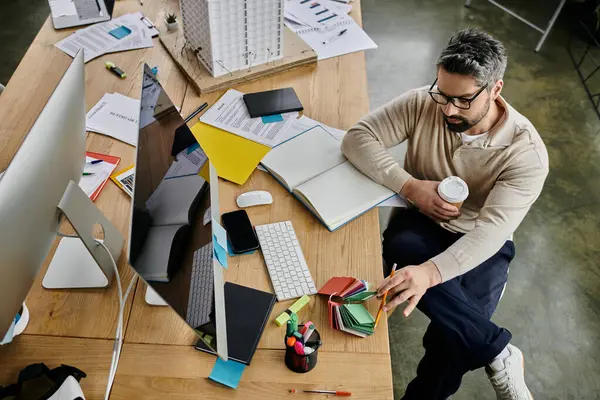 Um belo homem de negócios com barba se senta em uma mesa desordenada em um escritório moderno, cercado por desordem e materiais de trabalho. — Fotografia de Stock