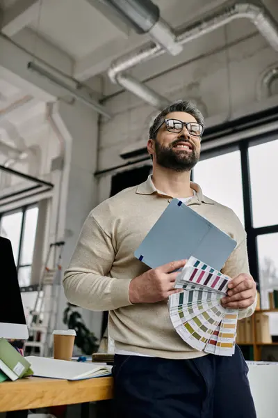 Un hombre de negocios barbudo, vestido con un suéter beige y gafas, se apoya contra un escritorio en una oficina moderna y sostiene muestras de color y una carpeta. — Stock Photo