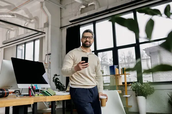 Um belo homem de negócios com barba trabalha em um escritório moderno, segurando um smartphone e uma xícara de café. — Fotografia de Stock