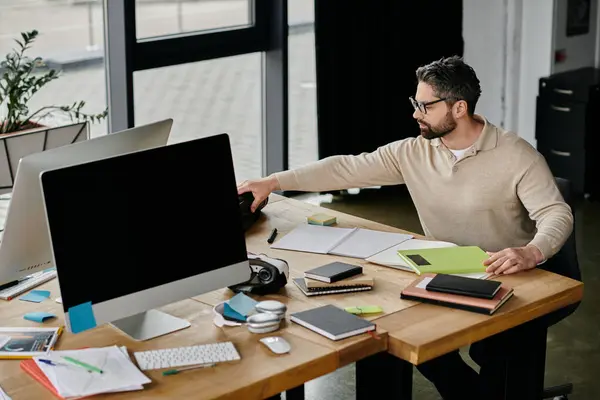 Un bel homme d'affaires avec une barbe travaille à son bureau dans un bureau moderne, avec deux moniteurs et plusieurs cahiers et stylos. — Photo de stock