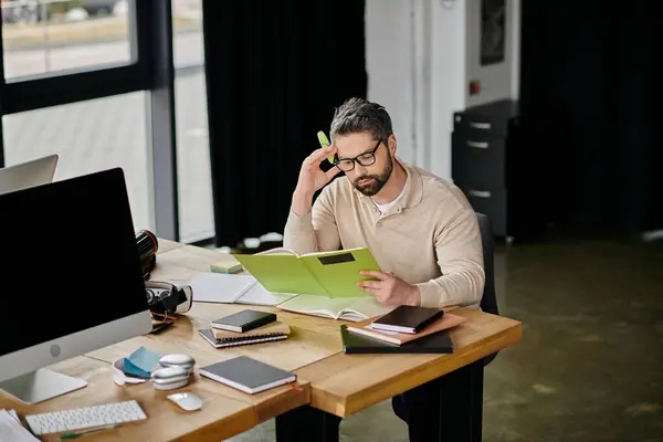 Un bell'uomo d'affari con la barba si siede a una scrivania in un ufficio moderno, leggendo intensamente un libro. Indossa occhiali e ha un'espressione premurosa. — Foto stock