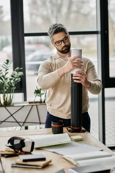 Un hombre de negocios guapo con barba cuidadosamente coloca un plano en un tubo mientras está de pie en su oficina moderna. - foto de stock