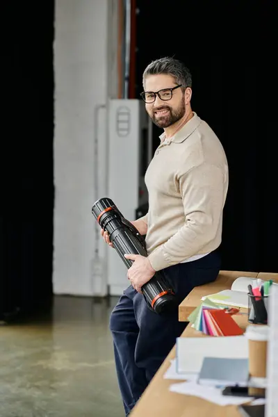 Un bell'uomo d'affari con la barba si siede su una scrivania moderna, tenendo un tubo e sorridendo con fiducia. — Foto stock