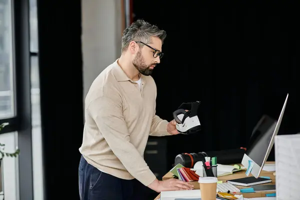 A handsome businessman with a beard examines a virtual reality headset while working in his modern office. — Stock Photo