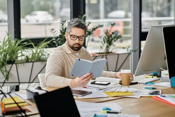 Un bell'uomo d'affari con la barba si siede ad una scrivania in un ufficio moderno, leggendo un libro. — Foto stock