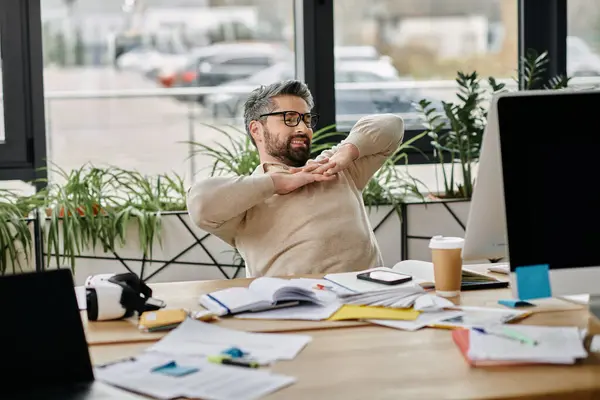 A businessman with a beard stretches at his desk in a modern office setting. — Stock Photo