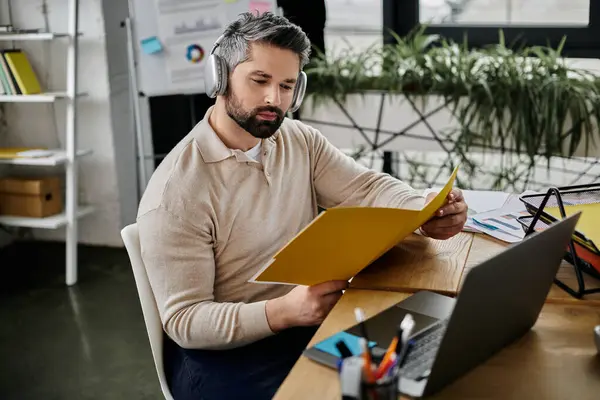 A handsome businessman with a beard is focused on reviewing documents in a modern office setting. He is wearing headphones, suggesting he is listening to music or working on a project that requires concentration. — Stock Photo