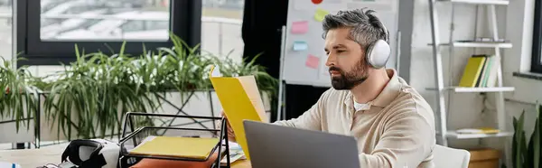 A handsome businessman with a beard is working at his desk in a modern office, wearing headphones and reviewing a folder. — Stock Photo