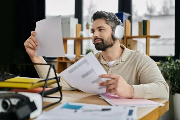 A handsome businessman with a beard sits at his desk in a modern office, reviewing documents while wearing headphones. — Stock Photo