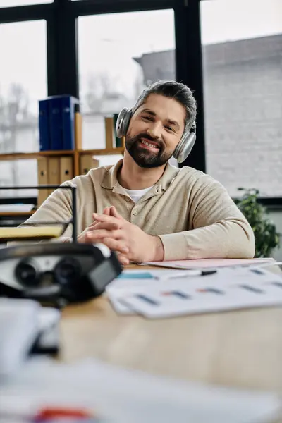A handsome businessman with a beard smiles while listening to music through headphones at his desk in a modern office. — Stock Photo