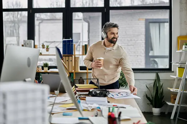 A bearded businessman in headphones stands at a messy desk in a modern office, holding a coffee cup and looking away from the camera. — Stock Photo