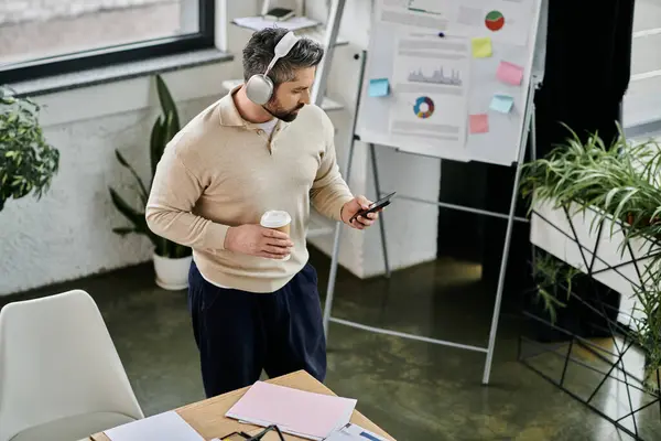 Um empresário barbudo em uma camisola bege fica em um escritório moderno, segurando uma xícara de café e verificando seu telefone. — Fotografia de Stock
