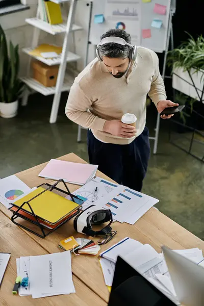 A handsome businessman with a beard takes a break in a modern office, holding a cup of coffee and checking his phone. — Stock Photo
