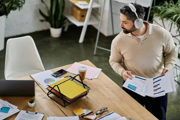 Un hombre de negocios guapo con barba está revisando cartas y gráficos en su escritorio de oficina moderno. Lleva auriculares y parece concentrado en su trabajo.. — Stock Photo
