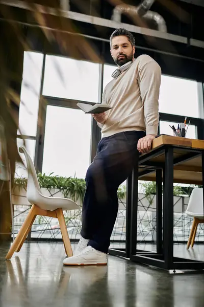 A handsome businessman with a beard leans against a modern office desk while holding a book, looking confident and focused. — Stock Photo