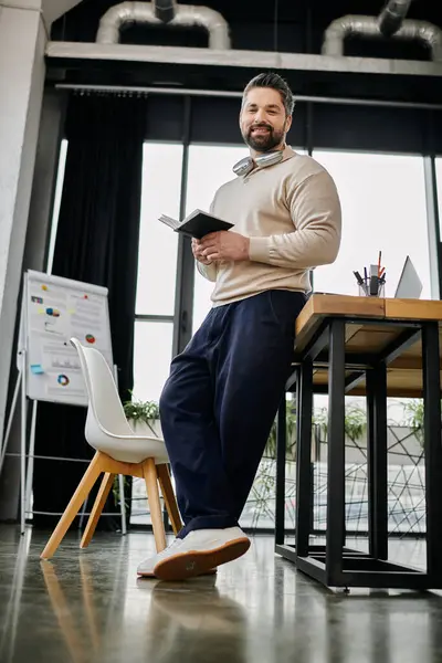 A handsome businessman with a beard is standing confidently in a modern office, leaning against a desk while holding a notebook. — Stock Photo