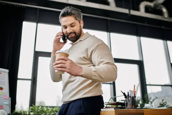 A handsome bearded businessman stands in his modern office, talking on his phone while holding a cup of coffee. — Stock Photo