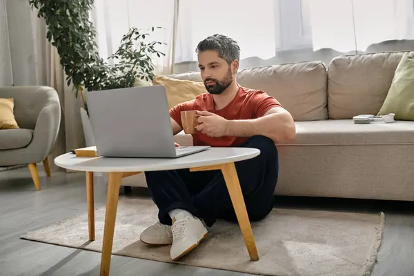 Un homme barbu en tenue décontractée est assis sur le sol devant une table avec un ordinateur portable, travaillant à distance de la maison. — Photo de stock