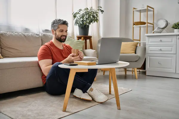 Un hombre barbudo con atuendo casual se sienta en el suelo, trabajando en su computadora portátil en casa. - foto de stock